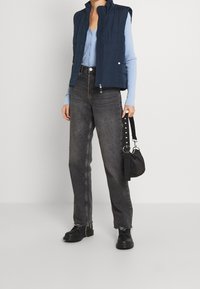 Navy padded vest with a high collar, paired with a light blue ribbed shirt and black high-waisted jeans. Black handbag with a strap.