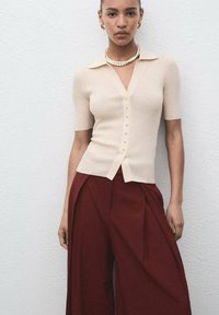 Vrouw in een beige, kort mouwige, gebuttonede top, maroon wijde broek, gouden creolen en een parelketting, staande tegen een witte muur.