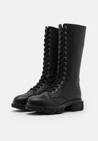 Bottes de combat noires en cuir, montantes jusqu'au genou, avec une semelle en caoutchouc épaisse, présentant des œillets en métal et des détails de laçage complexes à l'avant.