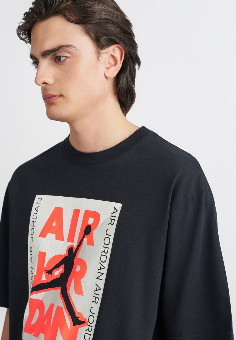 Μαύρη βαμβακερή μπλούζα με μεγάλο γραφικό σχέδιο που περιλαμβάνει το κείμενο "AIR JORDAN" και τη σιλουέτα ενός παίκτη που κάνει σουτ με κόκκινο και μαύρο χρώμα.