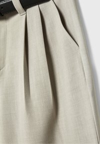 Gros plan sur un pantalon plissé beige clair avec poche latérale avant et passant de ceinture sur un cintre noir.