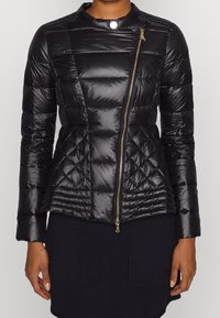 Czarna kurtka puchowa o dopasowanym kroju, pikowanym wykończeniu, wysokim kołnierzu i diagonalnym złotym zamkiem błyskawicznym. Charakteryzuje się gładkim, połyskującym materiałem.