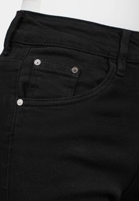 Jeans en denim noir avec un design classique à cinq poches, quincaillerie en métal argenté et un tissu texturé avec des détails de couture visibles.