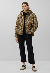 Leopardprint pufferjack met een capuchon, zwarte rits en gestructureerde stof; gecombineerd met zwarte jeans en crèmekleurige sneakers met bruine accenten.