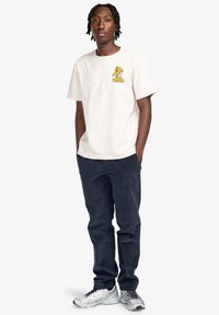 Camiseta de algodón beige claro con un diseño gráfico amarillo en el pecho, combinada con pantalones de pana oscuramente azul marino y zapatillas deportivas plateadas.