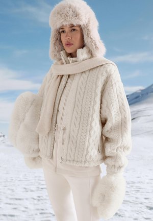 Fehér szőrmével bélelt sapka, krémszínű, texturált, fonott mintás kabát, hozzáillő sál és kesztyűk, világos krémszínű felsőre és szűk szabású nadrágra rétegezve.