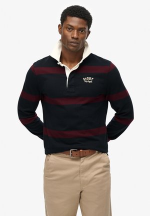 Man wearing a navy rugby shirt with maroon stripes and beige pants, standing with hands behind his back against a plain background.