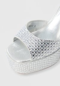 Srebrn podplat z visokimi petami in odprtimi prsti, okrašen z bleščečimi kristalčki in gladko kovinsko površino. Podplat z teksturo in bleščečimi poudarki.