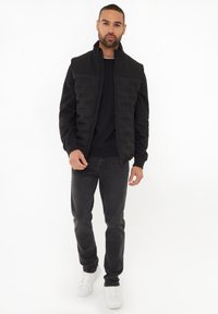 Schwarze Zip-Weste mit gepolsterter Vorderseite, kombiniert mit einem schwarzen Sweatshirt und dunklen Jeans. Weiße Sneakers runden das Outfit ab. Schlichter Hintergrund.