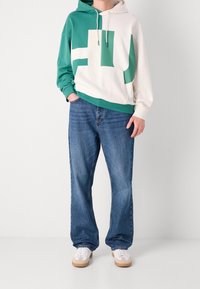 Zelená a krémová mikina s kapucí a geometrickými vzory, kombinovaná s modrými džínami a bílými teniskami s béžovými akcenty.