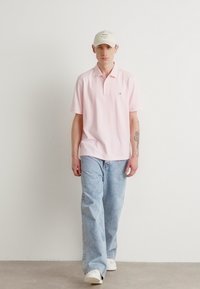 Polo shirt rose clair avec un col, associé à un jean en denim bleu clair et des baskets blanches. Une casquette beige complète la tenue.