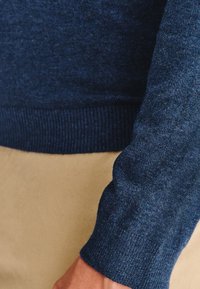 Zbliżenie na osobę w niebieskim swetrze z dzianiny i beżowych spodniach, skupiając się na teksturze materiału i kolorach.
