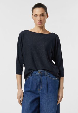 Woman with dark pulled-back hair wearing a loose dark navy three-quarter sleeve top and high-waisted blue jeans against a plain background.