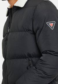 Veste matelassée noire avec col en polaire blanche, fermeture éclair frontale, poche latérale zippée et écusson triangulaire sur la partie supérieure de la manche avec une feuille d'érable rouge.