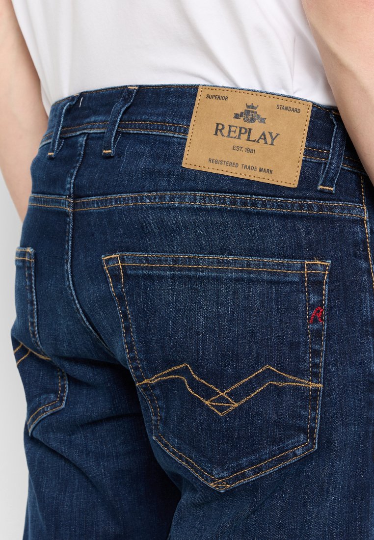 Jeans in denim blu scuro con un'etichetta in pelle contrassegnata "REPLAY". Presentano cuciture gialle a contrasto e un logo rosso sulla tasca posteriore.