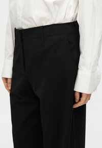 Pantalon noir sur mesure à coupe droite, avec passants de ceinture, poches latérales et une texture lisse. Port é avec une chemise blanche à boutons.