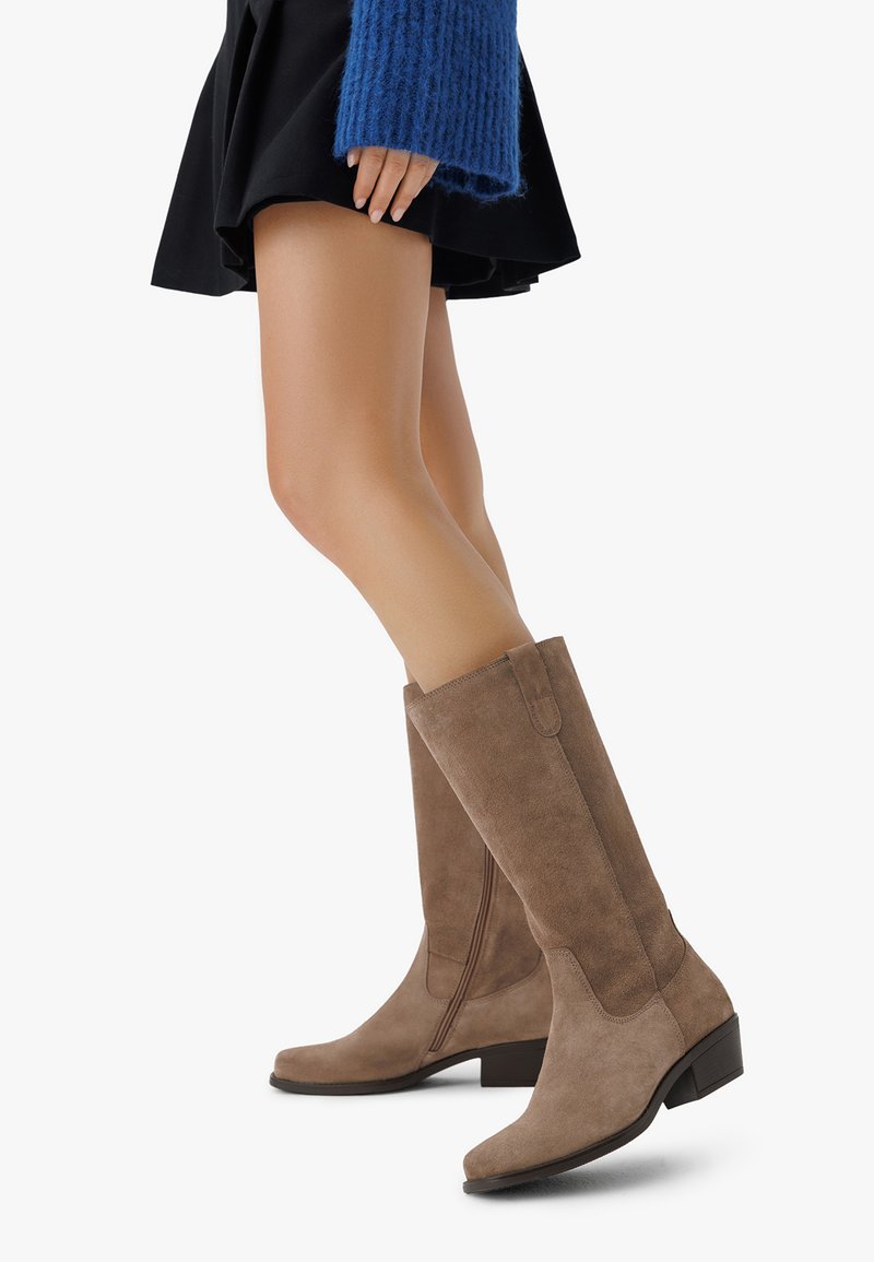 Botas de ante marrón hasta la rodilla con punta cuadrada, tacón bloque bajo y detalle de cremallera lateral, exhibidas junto a una falda negra.