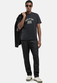 Camiseta negra con el texto blanco "REPLAY1981 DENIM GOODS", chaqueta negra con parches, pantalones oscuros, zapatos claros y gafas de sol.