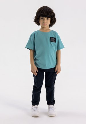 Turquoise katoenen T-shirt met korte mouwen, voorzien van een zwart labelpatch, gecombineerd met donkerblauwe cargobroek en witte sneakers.