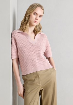 Mujer con cabello rubio que lleva un top de punto de manga corta color rosa claro y pantalones beige, apoyada en una pared blanca.