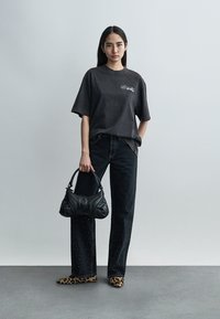 Γυναίκα που φοράει μαύρο t-shirt Off-White, μαύρο τζιν με τρουκς, παπούτσια με λεοπάρ σχέδιο, κρατώντας μαύρη τσάντα, στέκεται μπροστά σε απλό γκρι φόντο.