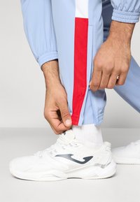 Ανοιχτό μπλε αθλητικό παντελόνι με κόκκινη λωρίδα στο πλάι, ελαστικές μανσέτες και λεπτομέρειες με φερμουάρ. Συνδυασμένο με λευκά αθλητικά παπούτσια με μαύρες λεπτομέρειες.