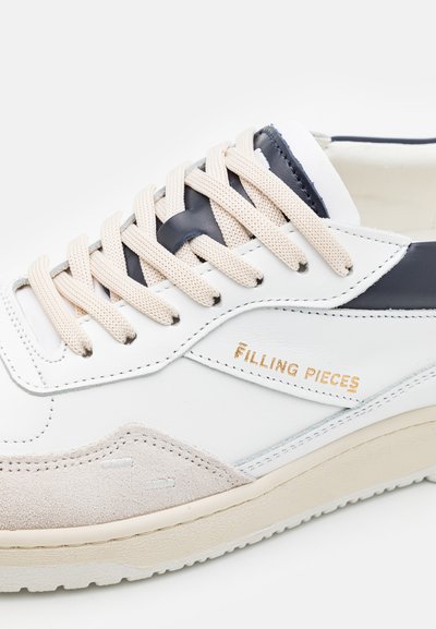 Bílé tenisky s akcentem z béžového semiše, tmavě modrými koženými detaily a světlými béžovými tkaničkami. Obsahují zlaté logo "FILLING PIECES" na boku.