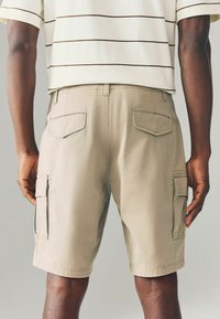 Beige cargo shorts, valmistettu puuvillasta, joissa on sivutaskut ja kaksi takataskua. Kangas on sileä ja siinä on ompelusarjat.
