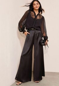 Schwarze, durchsichtige Bluse mit Spitzenverzierungen, kombiniert mit weit geschnittenen schwarzen Satinhosen und einer kleinen schwarzen Handtasche. Flache schwarze Sandalen runden den Look ab.