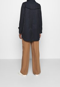 Persona vestida con un abrigo trinchera azul marino, pantalones de pierna ancha color camel y zapatillas blancas, de pie contra un fondo claro liso, vista desde atrás.