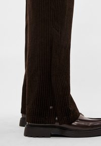 Brūnas kordurova bikses ar vertikālām ribām un sānu šķēlumiem apakšmalās. Kopā ar spīdīgām tumši brūnām ādas kurpēm.