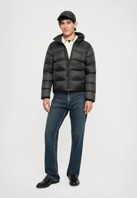 Schwarze Daunenjacke mit Kapuze, Reißverschluss und gestepptem Design. Kombiniert mit blauen Jeans und schwarzen Schuhen, stehend vor einem neutralen Hintergrund.