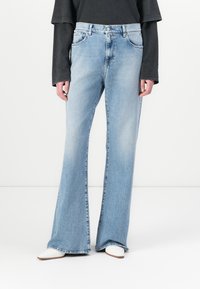 Blugi deschiși la culoare, evazați, din denim, cu talie înaltă, cinci buzunare și margini desfăcute, asortați cu un top negru cu mâneci lungi și încălțări albe.