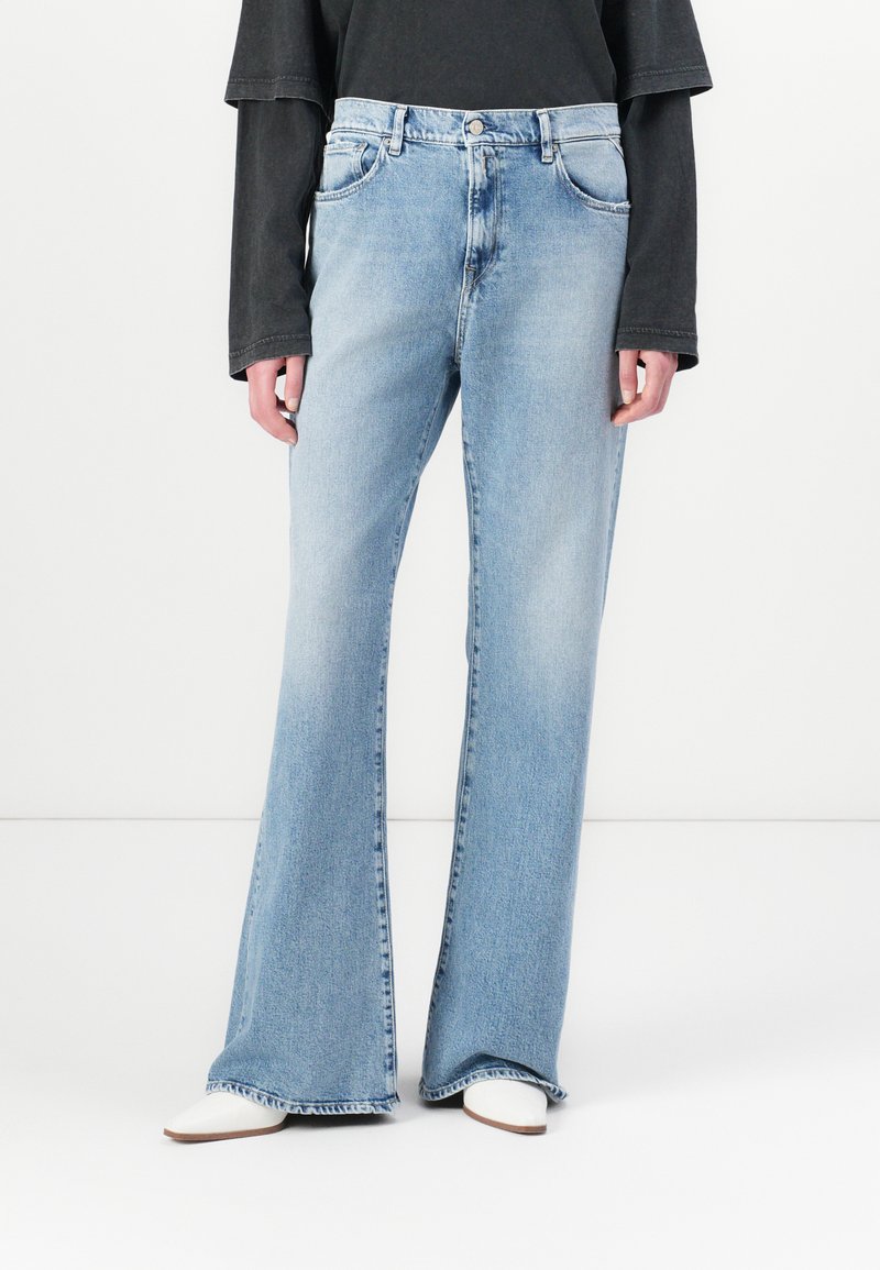 Blugi deschiși la culoare, evazați, din denim, cu talie înaltă, cinci buzunare și margini desfăcute, asortați cu un top negru cu mâneci lungi și încălțări albe.