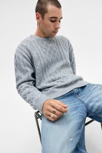 Lys grå kabelstrikket sweater med ribbede manchetter og kant, båret over blå slidte jeans. Model siddende på en metalstol, hænder synlige.