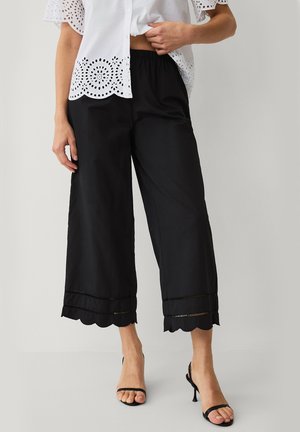 Kvinde iført sorte vidde, korte bukser med bølget kant, hvid broderet bluse og sorte åbne hælede sandaler, stående mod grå baggrund.