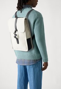 Kreemivärvi backpack, mis on valmistatud vastupidavast materjalist, flapi sulgemisega, mustade rihmade aktsentidega ja tekstuurse kudumisega sviiter all.
