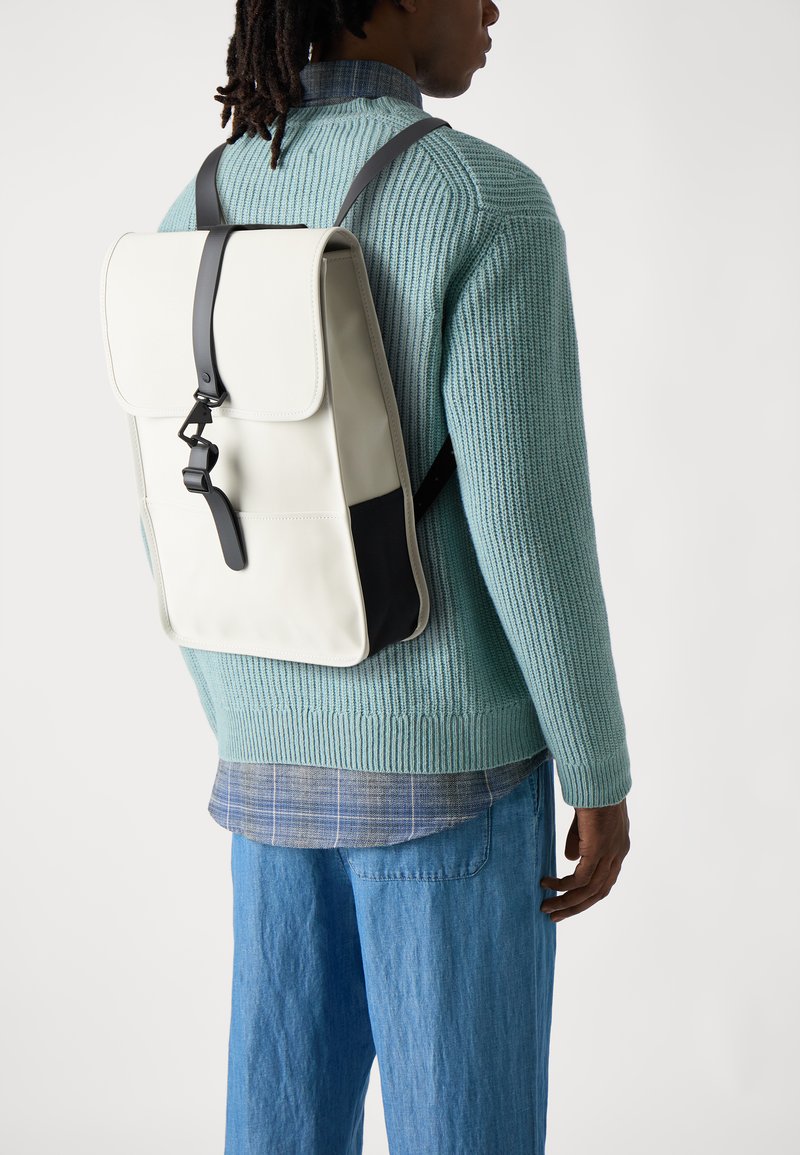 Un sac à dos de couleur crème en matériau résistant, présentant une fermeture à rabat, des accents de sangle noirs et un pull en tricot texturé en dessous.