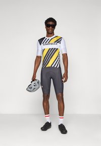 Sykkeldrakt med svarte og gule skråstriper, korte ermer, kombinert med grå shorts og svarte sko. Holder en grå hjelm.