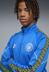 Sininen vetoketjulla varustettu takki, joka on valmistettu pehmeästä kankaasta ja jossa on Manchester City -logo, musta ja vihreä kuviointi hihoissa sekä valkoiset yksityiskohdat.