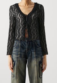 Svart spetsblus med vågig halsringning, långa ärmar och knytning framtill, kombinerad med löst sittande blå denimjeans.