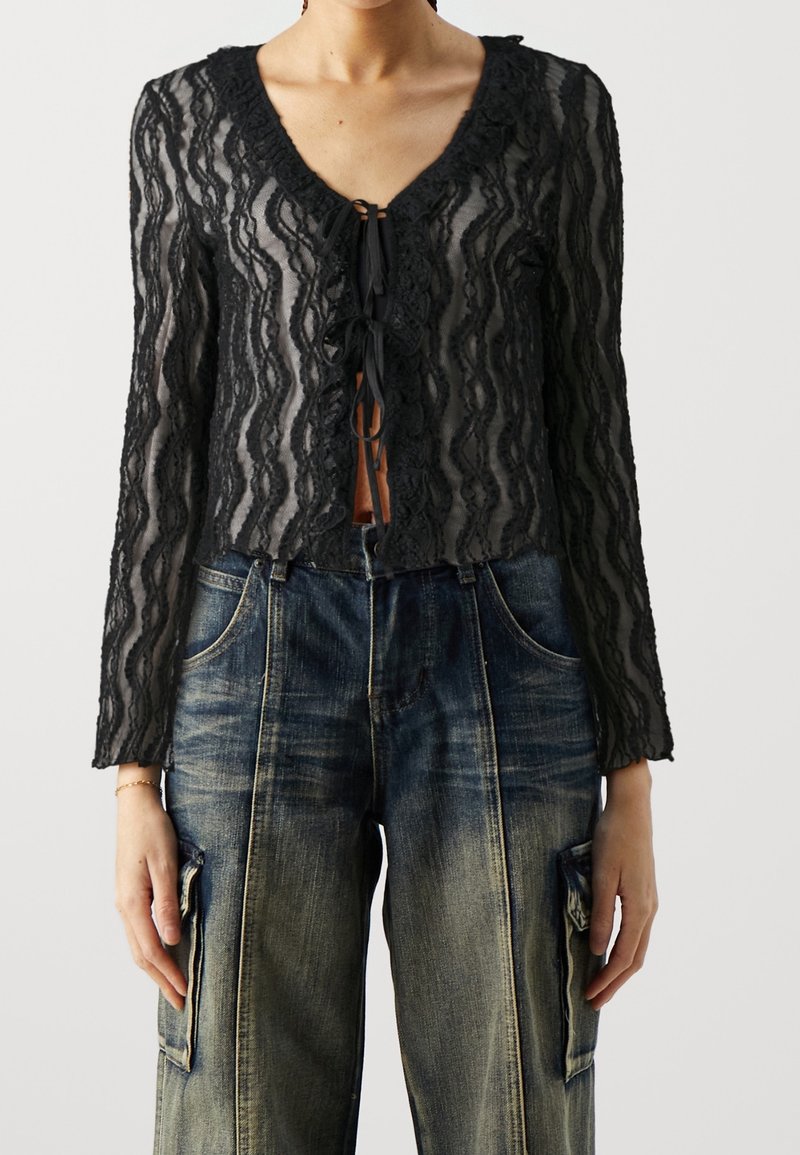 Svart spetsblus med vågig halsringning, långa ärmar och knytning framtill, kombinerad med löst sittande blå denimjeans.