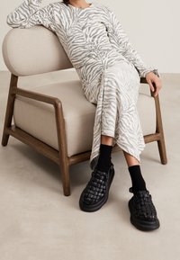 Tkaninowe czarne buty wsuwane z grubą podeszwą i teksturowanym wzorem, noszone w połączeniu z czarnymi skarpetami, zestawione z jasnoszarym sukienką w paski zebry i beżowym krzesłem.