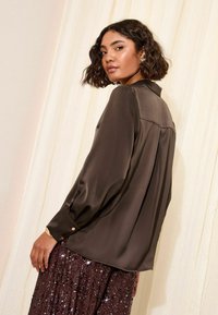 Bluza maro din satin cu manșetă cu nasturi, guler clasic și un luciu fin; asortată cu o fustă burgundy strălucitoare, cu paiete.