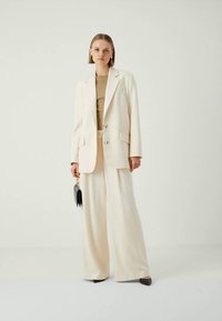 Elfenbenfarget skreddersydd blazer med to knapper, kombinert med vide bukser. Glatt stofftekstur. Modell som holder en liten svart veske.