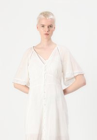 Robe blanche légère avec des manches courtes à volants, décolleté en V et boutons décoratifs sur le devant ; présente une bordure en dentelle sur les manches.