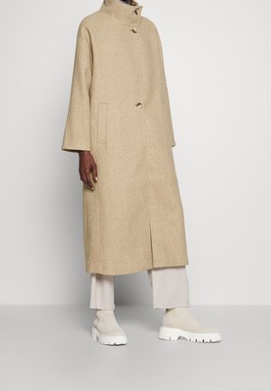 Długi beżowy płaszcz z wełny z wysokim kołnierzem i trzema dużymi guzikami, noszony na jasnobeżowe, szerokie spodnie i masywne białe botki.