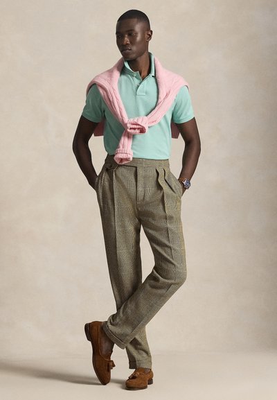 Άνδρας που φοράει ανοιχτό πράσινο πόλο, ροζ πουλόβερ ριγμένο στους ώμους, καρό παντελόνια, καφέ μοκασίνια και ρολόι χειρός, στέκεται με τα χέρια στις τσέπες.
