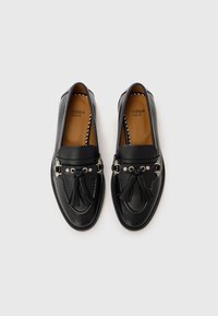 Une paire de mocassins noirs en cuir avec des pampilles et des œillets argentés, marqués "Toga Pulla" sur des semelles de couleur beige, sur fond blanc.