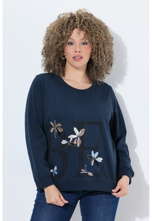 Frau mit lockigem Haar, die einen marineblauen Sweatshirt mit Blumenstickerei und dunkelblauen Jeans trägt und vor einem weißen Hintergrund steht.